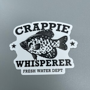 Op de afbeelding: Zwart-witte sticker met een baars en de tekst "Crappie Whisperer Fresh Water Dept."