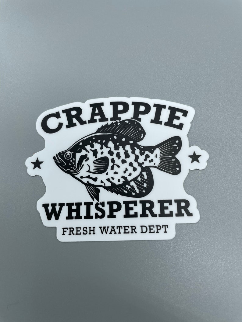 Op de afbeelding: Zwart-witte sticker met een baars en de tekst "Crappie Whisperer Fresh Water Dept."