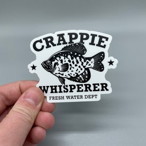 Op de afbeelding: Een witte sticker met een zwart-witte illustratie van een baars. De tekst "CRAPPIE WHISPERER FRESH WATER DEPT" staat op de sticker gedrukt.