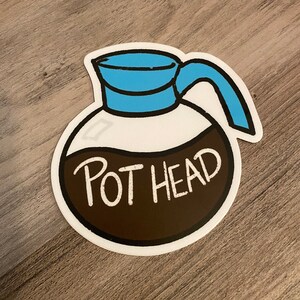 May include: A white sticker with a cartoon illustration of a coffee pot with a blue lid and handle. The coffee pot is filled with brown coffee and the text "POT HEAD" is written in white on the coffee pot.