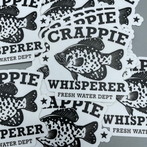 Op de afbeelding: Een stapel zwart-wit stickers met een visafbeelding en de tekst "Crappie Whisperer Fresh Water Dept" en "Appie Whisperer Fresh Water Dept".