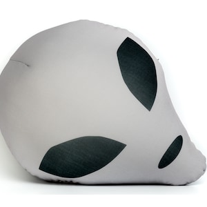 May include: A gray pillow with a black alien face design. The alien has two large eyes and a small mouth.