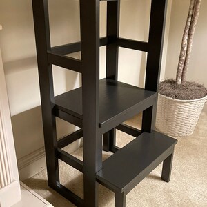 Puede incluir: Un taburete de madera negro con un estante incorporado. El taburete tiene tres pelda&ntilde;os y un estante superior. El taburete est&aacute; hecho de madera maciza y tiene un acabado liso.