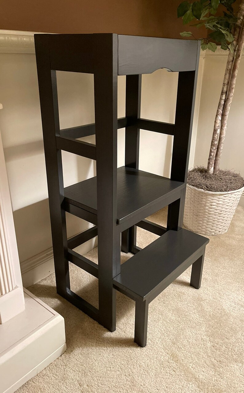 Puede incluir: Un taburete de madera negro con un estante incorporado. El taburete tiene tres pelda&ntilde;os y un estante superior. El taburete est&aacute; hecho de madera maciza y tiene un acabado liso.