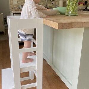 Puede incluir: Una torre de aprendizaje de madera blanca con un taburete de paso, que permite a un ni&ntilde;o alcanzar de forma segura una encimera de cocina. Un ni&ntilde;o peque&ntilde;o se para en la torre, alcanzando un taz&oacute;n en la encimera.