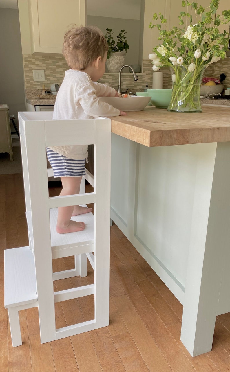 Puede incluir: Una torre de aprendizaje de madera blanca con un taburete de paso, que permite a un ni&ntilde;o alcanzar de forma segura una encimera de cocina. Un ni&ntilde;o peque&ntilde;o se para en la torre, alcanzando un taz&oacute;n en la encimera.