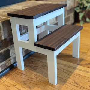 May include: A two-step stool featuring a white painted wooden frame and two dark brown stained wooden steps. The structure includes four vertical legs and horizontal supports connecting the upper and lower tiers in a simple, functional design.