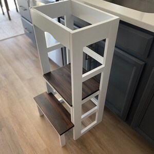 Puede incluir: Taburete de madera blanco con tres pelda&ntilde;os y una parte superior de madera marr&oacute;n. El taburete est&aacute; en una cocina junto a un fregadero.