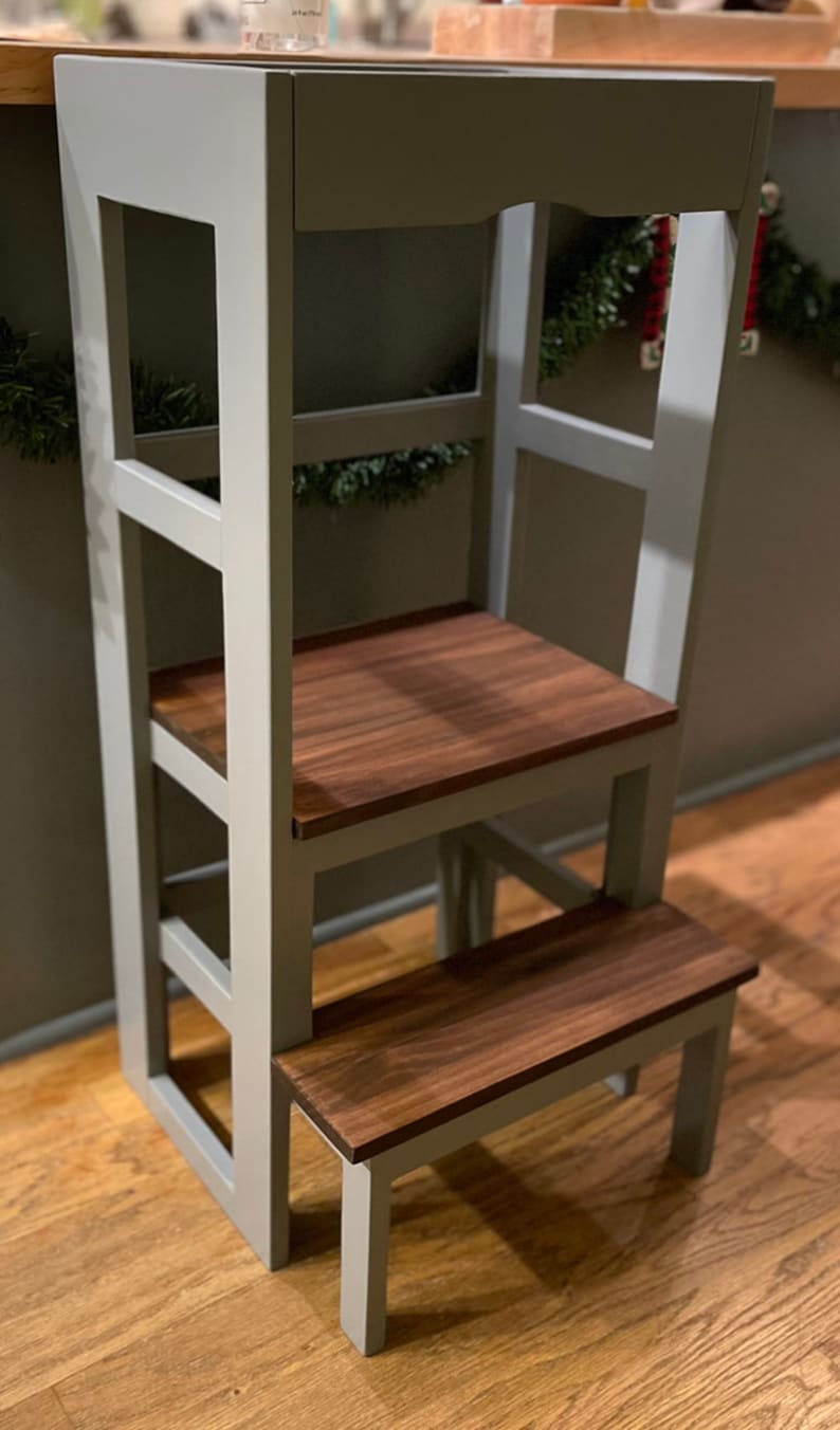 Puede incluir: Un taburete de madera gris con dos pelda&ntilde;os y una plataforma superior. Los pelda&ntilde;os y la plataforma son de madera oscura. El taburete est&aacute; sobre un suelo de madera.
