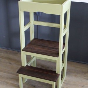 Puede incluir: Un taburete de madera de color verde claro con una parte superior de madera marr&oacute;n oscuro y un escal&oacute;n inferior. El taburete tiene un dise&ntilde;o sencillo con un marco rectangular y un solo estante.