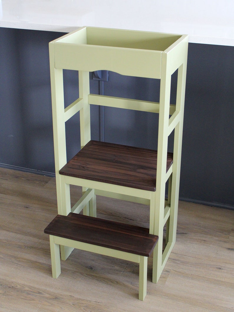 Puede incluir: Un taburete de madera de color verde claro con una parte superior de madera marr&oacute;n oscuro y un escal&oacute;n inferior. El taburete tiene un dise&ntilde;o sencillo con un marco rectangular y un solo estante.