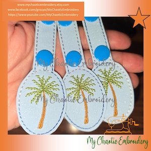 May include: Light blue keychains with a palm tree design. Each keychain has a light blue oval with a stitched palm tree in green and brown. The keychains have a blue button and a strap. The text "My Chaotic Embroidery" is visible.