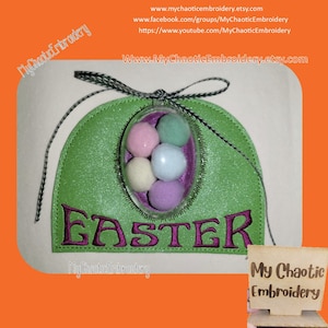 May include: Green felt Easter egg holder with the word "Easter" embroidered in purple. The holder has a clear plastic egg with five colorful pom-poms inside. The holder is decorated with a black and white ribbon.