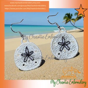 May include: A pair of white embroidered earrings in a sand dollar design with a black starfish pattern. The earrings have silver-colored hooks and are displayed on a sandy beach with a tropical background. The earrings are from My Chaotic Embroidery.