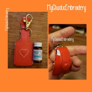 May include: Red leather keychain with a heart design and a small bottle holder. The bottle holder is designed to hold a small bottle of medication. The keychain is attached to a silver key ring.
