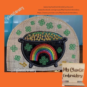 Puede incluir: Una olla negra llena de monedas de oro y un arcoíris se encuentra sobre un fondo de tela beige con tréboles verdes. El texto "My Chaotic Embroidery" está en un letrero de madera.
