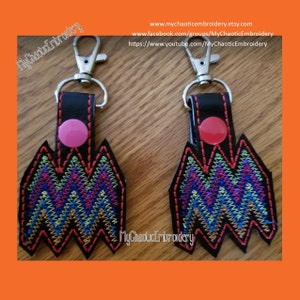 Puede incluir: Dos llaveros de cuero negro con anillas de llave plateadas. Cada llavero presenta un diseño bordado en rojo, negro, azul, verde y amarillo de una cordillera estilizada. Los llaveros tienen un botón rosa y rojo en la parte superior.