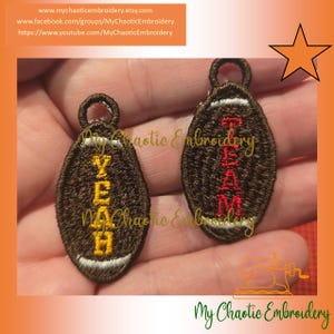 May include: Two embroidered football-shaped pendants. One says "YEAH" in yellow, the other says "TEAM" in red. Both are brown with white and yellow accents. The pendants have a loop at the top for hanging.