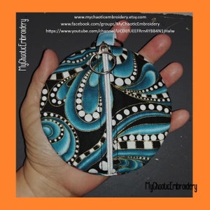 May include: A round zippered pouch with a black background and a blue and white paisley pattern. The pouch has a gold zipper and a silver ring attached to the zipper pull. The pouch is being held in a hand.