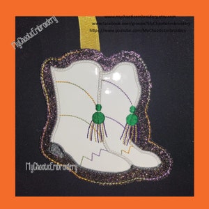 Puede incluir: Adorno en forma de bota de vaquero blanca con ribete de purpurina morada y cinta dorada. Las botas tienen detalles de borlas verdes, doradas y moradas. El texto "MyChaoticEmbroidery" es visible.