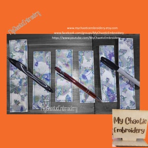 Puede incluir: Cinco marcapáginas de cuero blanco con un patrón de mariposa azul y morado. Cada marcapáginas tiene un bolígrafo o marcador negro en la parte superior. Los marcapáginas están sobre una superficie de madera. El texto "My Chaotic Embroidery" es visible en la esquina inferior derecha.