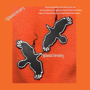 May include: A pair of black crow earrings with white stitching. The earrings are on an orange background.