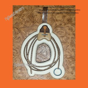 May include: A white embroidered design of a lasso and saddle on a brown leather background. The design includes the text "My Chaotic Embroidery" and a website address.