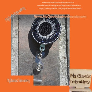 May include: A black and white eye-shaped embroidered badge with glitter accents attached to a retractable badge holder. The badge is on a gray textured surface. The text "My Chaotic Embroidery" is visible in the image.