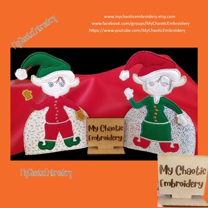 May include: Two embroidered Christmas elves, one in red and green, the other in green and white, stand on a red background with a wooden sign that reads "My Chaotic Embroidery".