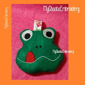 May include: Green felt frog with embroidered eyes, a red tongue sticking out, and a white smile. The frog is on a pink background with the text "MyChaoticEmbroidery" in the top right corner.