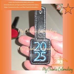 May include: Black leatherette keychain with the numbers "2025" embroidered in blue. The keychain has a rectangular tag with rounded corners and a strap for attachment. The text "My Chaotic Embroidery" is also visible.