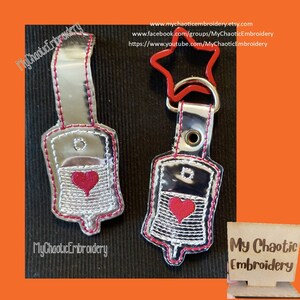 Puede incluir: Dos llaveros metálicos plateados con forma de bolsas de suero con corazones rojos. Los llaveros están unidos a anillos plateados con un llavero en forma de estrella roja en el llavero derecho. El texto "My Chaotic Embroidery" está en la parte inferior izquierda y derecha de la imagen.