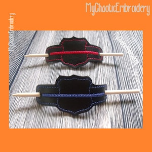 May include: Two black leather hair stick holders with a thin red line and a thin blue line, respectively. The holders are shaped like a shield with a stitched border. The sticks are white.