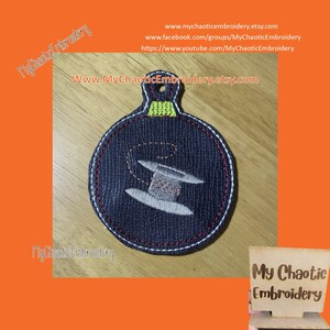 May include: A blue and white embroidered ornament shaped like a spool of thread. The ornament has a red and white stitched border. The ornament is on a wooden surface.