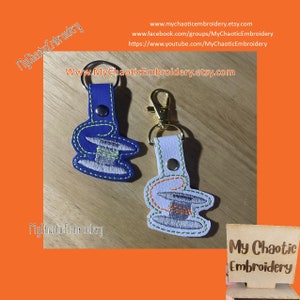 May include: Two embroidered keychains featuring a spool of thread. The keychain on the left is blue with green stitching. The keychain on the right is white with orange and green stitching. Both keychains have a gold metal ring.
