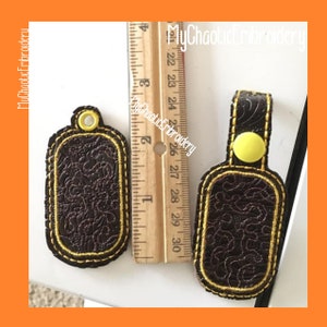 May include: Two brown leather keychains with yellow stitching. One keychain has a yellow button and the other has a ring. Both keychains have a decorative embossed pattern.