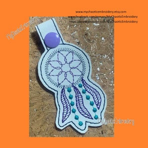 Könnte beinhalten: Weißer Filz-Traumfänger-Schlüsselanhänger mit lila und türkisfarbener Stickerei. Der Traumfänger hat ein kreisförmiges Design mit einer Blume in der Mitte und herabhängenden Federn. Der Schlüsselanhänger hat einen lilafarbenen Knopf oben.