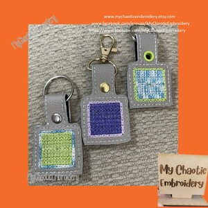 Puede incluir: Tres llaveros de cuero gris con cuadrados bordados de diferentes colores. Los llaveros están unidos a anillas de llaves doradas. Los llaveros están sobre una superficie gris texturizada. El texto "My Chaotic Embroidery" es visible en un letrero de madera en la esquina inferior derecha de la imagen.