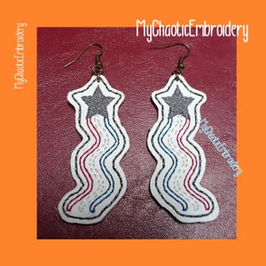 May include: A pair of white leather earrings with a star and red, white, and blue stripes. The earrings are shaped like a wavy line and have a silver hook.