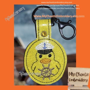 May include: Yellow glitter keychain with a cartoon yellow duck wearing a blue and white sailor hat and holding a brown steering wheel. The keychain has a black metal clasp.