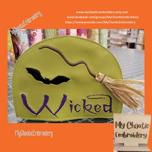 May include: A green felt witch hat with a purple embroidered "Wicked" text, a black bat, and a brown broom with a straw broom head. The hat is on a red surface with a wooden sign that says "My Chaotic Embroidery".