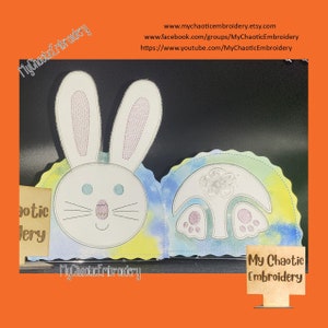 May include: A white felt bunny and mushroom applique with a tie-dye background. The bunny has blue eyes and a pink nose. The mushroom has a white top and a pink bottom. The text "My Chaotic Embroidery" is on a brown sign.