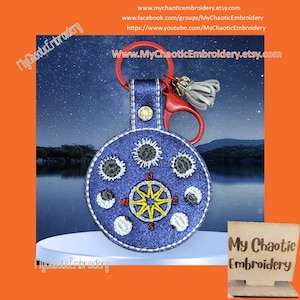 May include: A blue leather keychain with a sun and moon design. The keychain features a compass with the letters N, S, E, and W. The text "My Chaotic Embroidery" is visible on the keychain and on a wooden sign.