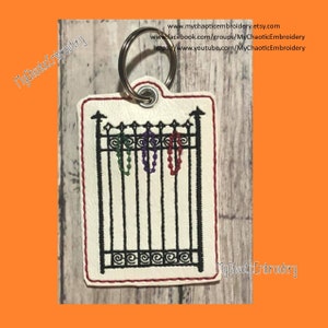 May include: A white leather keychain with a black embroidered image of a gate with three strands of colorful beads hanging from it. The text "MyChaoticEmbroidery" is on the keychain.