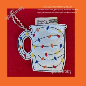 May include: White mug with a string of colorful lights around it. The mug has a handle and is decorated with a sparkly finish. A $50 bill is tucked into the top of the mug.