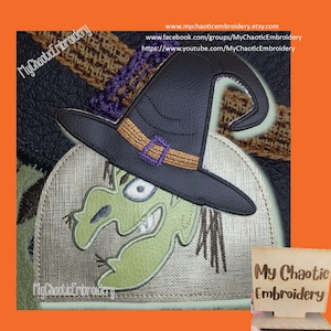 May include: Green witch face with a black witch hat and orange and purple trim. The witch face is embroidered on a beige fabric with the text "My Chaotic Embroidery" in the bottom left corner. The text "My Chaotic Embroidery" is also on a wooden sign in the bottom right corner.