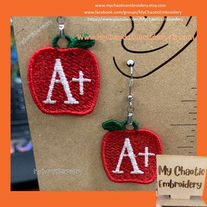以下が含まれることがあります： 赤いリンゴの形をしたイヤリングのペア。それぞれに白い「A+」が刺繍されています。イヤリングは銀色のフックからぶら下がっています。