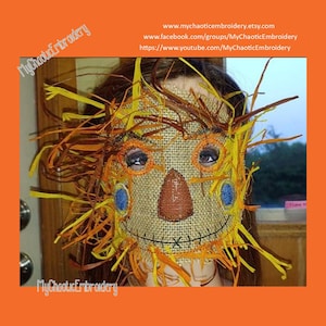 May include: A close-up of a scarecrow mask made of burlap with a stitched smile, orange eyes, and a brown nose. The mask is surrounded by yellow and orange straw-like material.
