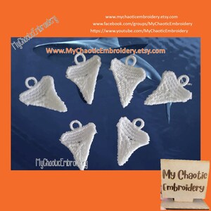 May include: Six white embroidered shark tooth charms with a loop for hanging. The charms are on a blue background. The text "My Chaotic Embroidery" is visible in the image.