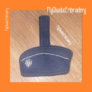 May include: A blue fabric key chain with a white embroidered star and a loop for attaching keys. The key chain is shaped like a hat with a brim and a top.